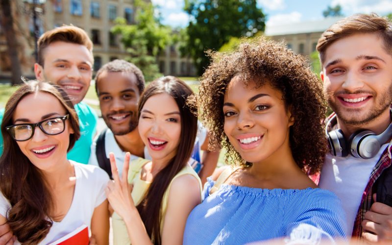Selfie time! Six international students with beaming smiles are posing for selfie shot, african attractive lady is taking, outside school building. Gathered, cheerful, smart and successful youth